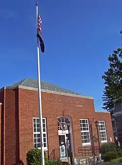 Three-quarter angle view of post office, a small brick building with hipped roof and a flagpole in front