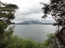 Image showing a still Lake Rotomahana on an overcast day framed by trees. Mount Tarawera is visible in the background