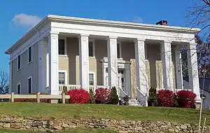 A two-story light brown wooden house with a flat roof and square columns in front on a rise in front of the camera