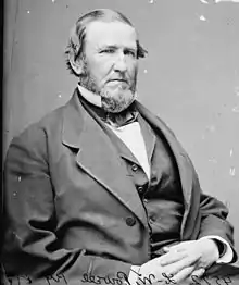 A man in his late forties with receding hair and a full beard. He is wearing a white shirt with a black vest and jacket and is facing right