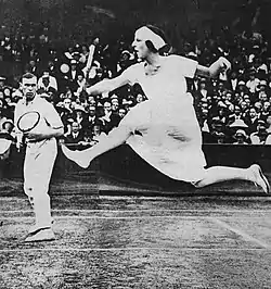 Lenglen hitting her signature leaping volley shot at the net from the advantage side of the court, with her partner Gerald Patterson watching at the net on the deuce side of the court