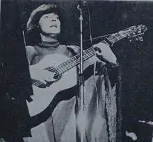  Black and white photograph of a short-haired woman in a poncho standing behind a microphone, playing a guitar.