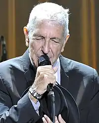 A man holding a guitar, wearing a blue shirt and a dark vest