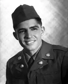 Head and shoulders of a smiling young man with dark hair wearing a garrison cap and a military jacket with two round pins on each lapel.