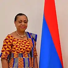 An African woman in traditional dress stands beside a red and blue flag