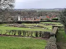 Low reddish-coloured stone walls, the construction of which suggests antiquity, lie in a grassy area backed by the outline of a modern settlement including a church with a spire. The trees lack leaves and the sky is a clear blue.