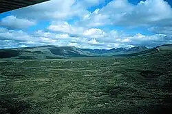 A broad, low and gently sloping mountain cut by a U-shaped valley rising over a vegetated plain.