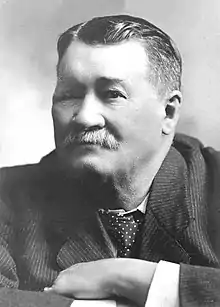 A headshot of a white man with a white moustache