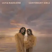 Lily and Madeleine standing side by side wearing frilly yellow and beige dresses in front of a cloudy sky over water