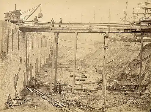 Enlargement (top-hatted directors posing, early 1867).  Note sailing ships — held back by temporary earth dam.