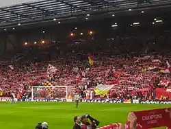 A single tiered stand that contains thousands of people. Several flags are being waved. In front of the stand is a grass pitch with a goal.