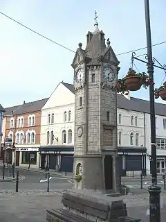 A tower-like structure in stone with a clock face on each side.  Above each clock is a gable and at the summit of the structure is a spire.