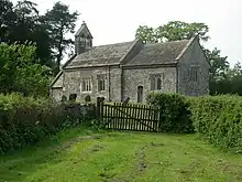 A small stone church seen from an angle; behind the chancel is a slightly higher nave with a bellcote at the far end. A porch protrudes beyond that