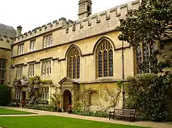 Adjoining buildings with battlements to the side of an area of grass; on the left, regularly-placed windows in a three-storey building, and foliage and flowers growing above and around a stone canopy over the doorway; on the right, a chapel with three arched windows and a stone porch; a bench alongside the chapel, and a tree at the far right