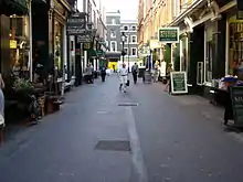  Narrow street with shops on each side. A few people are walking or window-shopping.