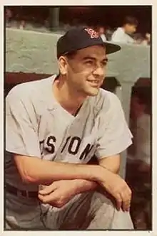 A man in a grey baseball uniform and blue hat leans out of a dugout.