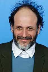 A middle-aged Caucasian man with receding short black hair, a short graying beard, and a mustache stands before a blue background with white text. He wears a tweed jacket, a blue sweater, and a collared, white shirt.