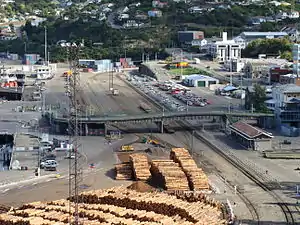 Lyttelton railway station yard. Features of interest include the Oxford Street overbridge (middle), Lyttelton station building (right, in front of the bridge), shore-end of wharves 2 and 3 (left, opposite the end of the bridge), Lyttelton rail tunnel portal (obscured, centre background), and Lyttelton road tunnel portal (centre-right background). Also shown are two of the ports main sources of traffic: timber and motor vehicles.