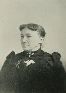 B&W portrait photo of a middle aged woman with her hair in an up-do, wearing a dark, high-collared blouse, her spectacles pinned to it.