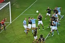 An action shot of two competing football teams trying to win a flying ball in the penalty area of one of the teams, the ball is heading for the head of the attacking team's captain. The players of the attacking team are wearing sky-blue shirts, white shorts and sky-blue socks. The field players of the defending team are wearing black shirts, white shorts and black socks. The goalkeeper for the defending team is wearing an orange shirt, black shorts and black socks.