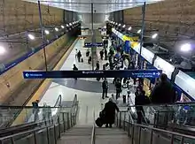 An underground center-platform railway station with sandy colored walls and people getting on and off a train on the right side of the image. Taken from the top of an escalator looking down.