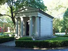 A brown stone building with a pointed roof and four columns in front in the middle of a paved circle and shrubbery. Across the top it says "Woodward".