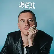 The cover features Macklemore against a blue background, wearing a black leather jacket, white t-shirt and gold rings on his left hand. The album title appears above him, colored in white.