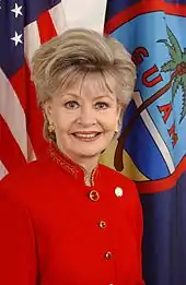 A blonde woman in red shown from the chest up standing in front of the flags of the United States and Guam