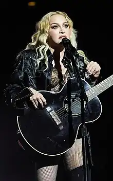 Madonna standing in front of a microphone, with a blank guitar strapped across her chest.