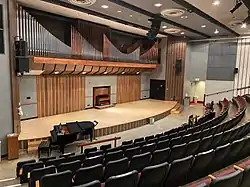 a small stage with a pipe organ behind and a piano on the floor in front. Sound equipment peers down from the ceiling, and rows of red chairs face the stage.