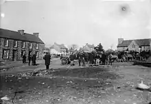 monochrome view of carts and people with houses and church beyond