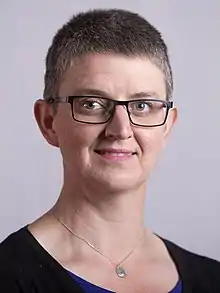 Official Portrait of Maggie Chapman MSP. She is smiling, with short black-ish hair and a necklace. There is a grey-ish background.