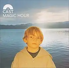 A young boy wearing a black t-shirt and grey hoodie with a sunlit lake behind him.