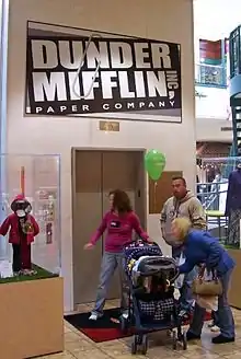 Three adults and a child in a stroller in front of a set of closed elevator doors. Above them is a sign with "Dunder Mifflin Paper Company" on it in white letters on a black background