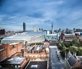 The image shows the Manchester Metropolitan University Business School, the All Saints campus and the Manchester skyline.