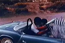 A man and woman kiss in a convertible sports car, with the clapperboard accidentally at the edge of the frame.