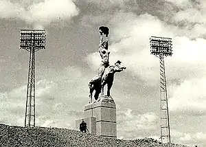 A monumental statue of a nude woman holding a pelvis and riding a tapir, standing between two floodlights.