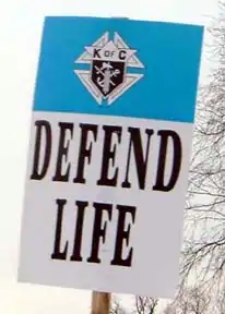 A photograph of a placard at the March of Life that reads "Defend life" on the bottom with the Emblem of the Order in a blue band on top.