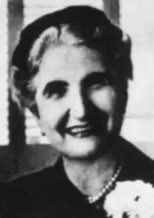 A smiling older white woman wearing a dark hat and suit with pearls and a corsage