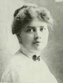 A young white woman, hair dressed in a bouffant updo, wearing a white shirt with a high collar and a small dark bow at the throat