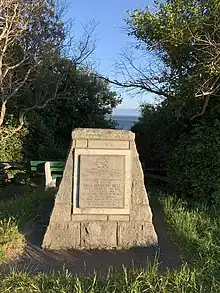 Stone cairn with engraved stone plaque depicting Marilyn Bell's portrait with swimming cap and goggles and waves in the background and the following text: This cairn commemorates the feat of Miss Marilyn Bell who landed in this bay 23rd August 1956 to become the first woman and first Canadian to swim Juan de Ducat Strait from Port Angeles USA to Victoria Canada.