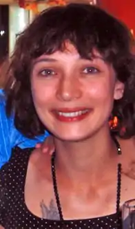 A smiling young woman, white with brown hair and green eyes, photographed indoors and illuminated by a flash. Behind her there is a person wearing a blue shirt whose face is not visible but has put a hand on the back of her neck. She is wearing a black beaded necklace and scoop-necked top; above her right breast two tattoed wings are visible