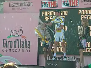 A cyclist in a yellow and white jersey with black trim popping a champagne bottle on a podium, flanked by two beautiful women holding flowers and wine.