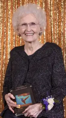 Martha Schwebach, a white lady in her eighties now, white-haired in a black dress, sits in a wheelchair holding her Nursing Legend Award in front of a gold curtain at an award ceremony. She smiles.