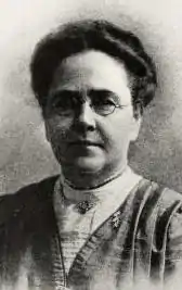 A middle-aged white woman wearing glasses and a high-necked blouse; her hair is arranged in a slightly bouffant top bun.