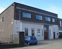 A brown-brick, flat-roofed, warehouse-style building. The lower half of its façade has been painted cream and decorated with arch-shaped window and door surrounds attached to the walls.  On the first floor there are three two-pane windows and one single window.  The side wall is mostly blank, with some small windows.