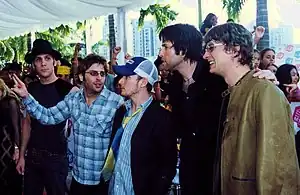 Five Caucasian men clad in casual attire pose together. In the background, a crowd has formed; several crowd members raise their arms.