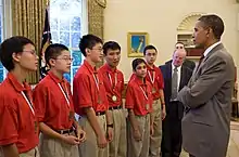 Six people in red shirts and beige pants standing in a line next to men in black suits all in front of a man wearing a grey suit and a red tie