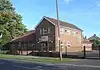 Three-quarter view of a vernacular-style brick building in two parts, behind a black fence and flanked by a grass area and a car park. The near part has two storeys with three windows above the entrance door and irregularly placed windows on the side wall.  The far section has a long, steep roof and one storey.