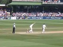 A bowler, midway through delivery.  An umpire with black trousers and a light shirt watches from the left while the batsman not receiving the delivery is already walking down the pitch.  In the background is a two-tier stand, full of spectators.